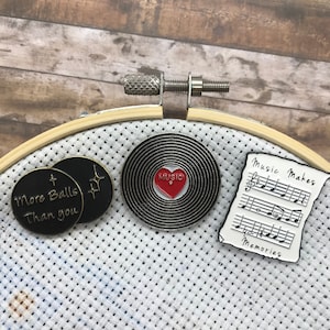 May include: Three enamel pins on a white fabric background. The pins are round and feature different designs. One pin is black with gold lettering that reads "More Balls Than You". Another pin is black with a red heart and the word "Music" in white lettering. The third pin is white with black musical notes and the text "Music Makes Memories".