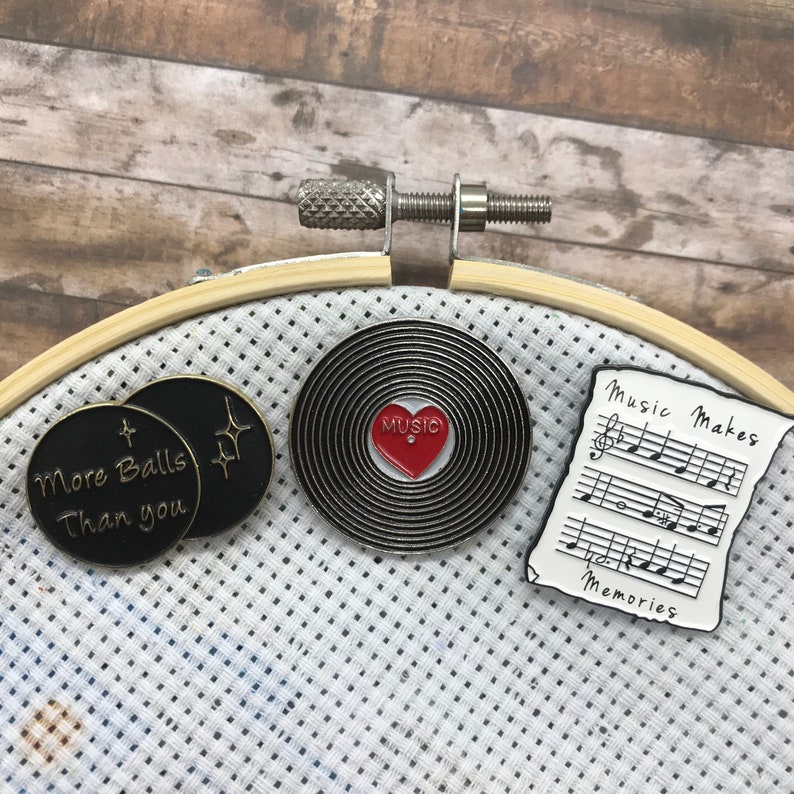 May include: Three enamel pins on a white fabric background. The pins are round and feature different designs. One pin is black with gold lettering that reads "More Balls Than You". Another pin is black with a red heart and the word "Music" in white lettering. The third pin is white with black musical notes and the text "Music Makes Memories".