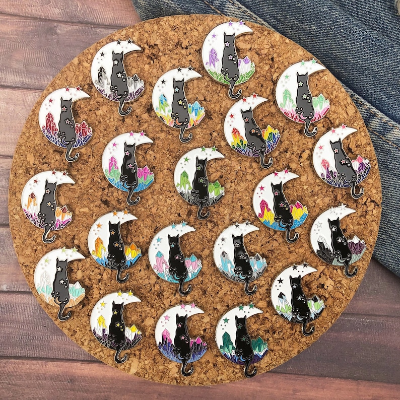 May include: Enamel pins featuring a black cat silhouette perched on a crescent moon with stars. Each pin has a different color crystal cluster at the base of the moon. The pins are arranged on a corkboard.