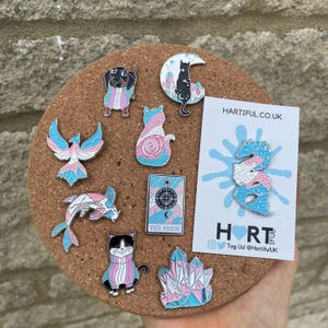 May include: A collection of enamel pins with a transgender pride theme. The pins feature various designs, including a dog, cat, phoenix, koi fish, tarot card, seahorse, and crystals, all in the colors of the transgender flag. The pins are displayed on a corkboard.