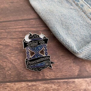 May include: A black and silver enamel pin with the text "Time To Procrastinate". The pin features an hourglass design with a starry blue interior, a crescent moon, and a star. The pin is on a wooden surface with a denim background.