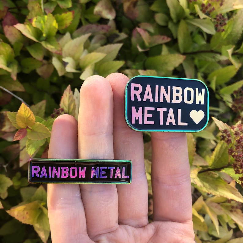 Peut inclure: Deux &eacute;pingles en &eacute;mail arc-en-ciel avec le texte "Rainbow Metal" sur fond noir. La plus grande &eacute;pingle a un c&oelig;ur rose. La plus petite &eacute;pingle est rectangulaire.