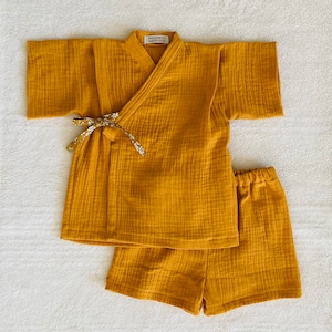 May include: Mustard yellow baby kimono-style top and shorts set. The top has a V-neck and a floral patterned tie closure. The shorts have an elastic waistband. The set is made of a textured fabric. The label reads "WASANBON handmade".