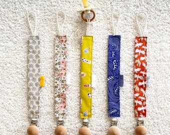 Könnte beinhalten: Fünf Baby-Schnullerclips mit verschiedenen Stoffmustern. Die Clips haben Holzperlen, Metallverschlüsse und Stoffbänder mit floralen, geometrischen und Wellenmustern. Ein Clip hält einen gelben Schnuller. Die Clips sind ca. 20 cm lang.
