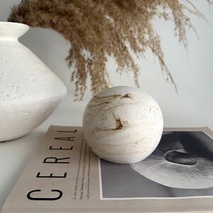 May include: A white ceramic sphere with brown marbling sits on top of a beige magazine with the word "CEREAL" printed on the cover. The magazine is on a white surface.