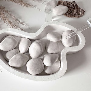 May include: White ceramic bowl with a unique shape, filled with smooth white stones. A glass dropper bottle and a small piece of driftwood are visible in the background.
