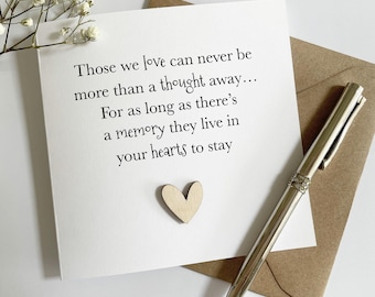 May include: A white greeting card with the text "Those we love can never be more than a thought away... For as long as there's a memory they live in your hearts to stay." A small wooden heart and a silver pen are also present.