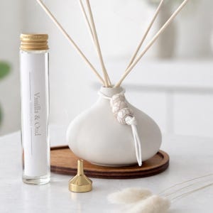 May include: A white ceramic diffuser vase with wooden reeds, a glass refill bottle labelled Vanilla & Oud with a gold cap, a small gold funnel, and two decorative beads on a white cord. The set is arranged on a dark wooden oval tray.