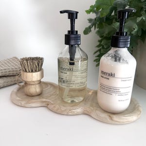 May include: A wooden, free-form tray holds two bottles of Meraki soap and lotion with black pumps, and a small wooden brush. The soap bottle is clear, and the lotion bottle is white. The tray is light brown.