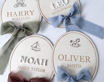 May include: Four oval-shaped wooden name plaques with engraved names and illustrations. Each plaque has a different color ribbon and features a name: Harry, Leo, Noah, and Oliver. The plaques have a teddy bear, train, sailboat, and rocking horse illustration.