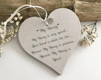Könnte beinhalten: Herzförmige Holzplakette mit dem Text "My Nanny" und einer liebevollen Botschaft. Die Plakette ist hellrosa mit einem dezenten Muster, und ein weißes Band ist zum Aufhängen befestigt. Kleine Blumen sind um die Plakette angeordnet.