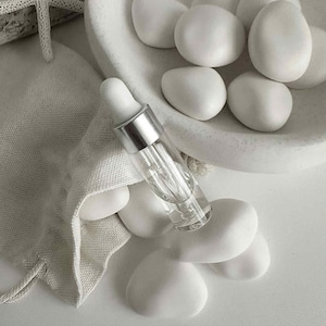 May include: A clear glass dropper bottle with a silver cap rests on white stones. A small white drawstring bag and a white starfish are also present. The items are arranged on a white surface, creating a minimalist aesthetic.