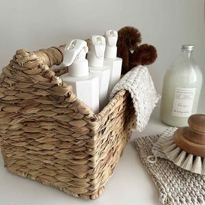 May include: A wicker basket contains white spray bottles. A glass bottle, a wooden brush, and a knitted washcloth are placed nearby. The items are arranged on a white surface, suggesting a cleaning or home care theme.