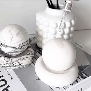 May include: Three white ceramic diffusers with black and white marble designs. The diffusers are on a book with the word "HAUS" printed on the cover. One diffuser has a glass dropper bottle with a clear liquid.