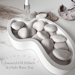 May include: White ceramic tray shaped like a wave with the text "Divine Home Essential Oil Pebbels & Onda Wave Tray". The tray is filled with white, round, speckled stones.