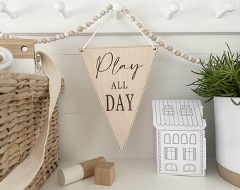 May include: A wooden pennant with the words "Play All Day" in a script font hangs from a white string. A woven basket, wooden toys, and a paper house are also visible, suggesting a children's room decor theme.