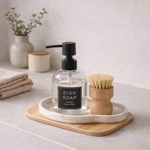 May include: A clear glass dish soap dispenser with a black pump and a label that reads "Dish Soap Clean Faactory." It sits on a marble tray with a wooden base, next to a wooden dish brush. The scene is set on a light-coloured countertop.