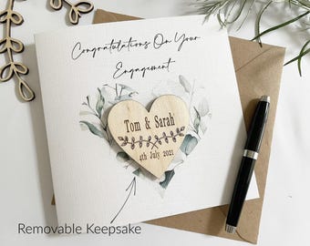 May include: A white engagement card with the text "Congratulations On Your Engagement". A wooden heart with the names "Tom & Sarah" and the date "4th July 2021" is attached. A black pen and a brown envelope are also present.