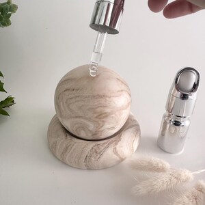 May include: A smooth, beige and brown marbled stone sphere resting on a matching circular base. A clear glass dropper with a silver metallic top is positioned above the sphere, releasing a single drop of clear liquid onto its surface.