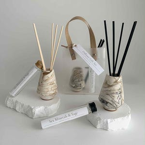 May include: A diffuser gift set with two ceramic vases, reed sticks, a clear bag, and glass tubes. The vases have a marbled beige and brown pattern. The tubes and bag are labelled "Sea Minerals & Sage". The set is designed for home fragrance.