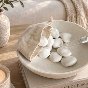 May include: A shallow, speckled beige ceramic bowl containing a small, light-coloured burlap drawstring pouch and several smooth, matte white stones. A clear glass dropper with a silver-toned metal collar rests inside the bowl among the stones.