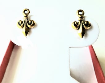 Könnte beinhalten: Ein Paar goldfarbene Ohrstecker, jeweils mit einem Fleur-de-Lis-Design. Die Ohrringe werden auf einem weißen Ohrringständer vor rotem Hintergrund präsentiert. Jeder Ohrring hat oben eine kleine Schlaufe.