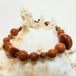 May include: A brown gemstone bracelet with gold accents. The bracelet is made of round and oval-shaped stones and is displayed on a white seashell.