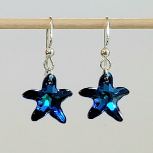 May include: A pair of silver dangle earrings with blue star-shaped crystal charms. The crystals have a dark blue color with a slight iridescent shimmer.