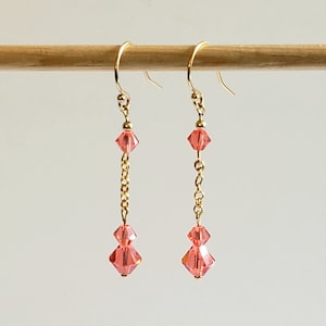 May include: A pair of gold dangle earrings with pink crystal beads. The earrings have a delicate chain design and feature two pink crystal beads on each earring.