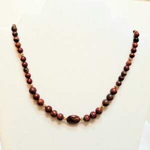 May include: A red, brown, and green stone beaded necklace with a gold accent. The necklace is made of round beads and has a larger oval-shaped bead in the center.