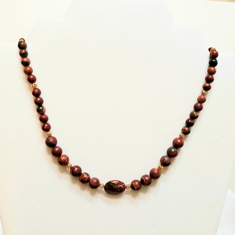 May include: A red, brown, and green stone beaded necklace with a gold accent. The necklace is made of round beads and has a larger oval-shaped bead in the center.