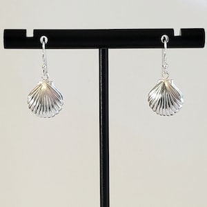 Op de afbeelding: Zilveren oorhangers met een schelpontwerp. De oorbellen bevinden zich op een zwarte standaard.