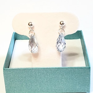 May include: A pair of silver-toned drop earrings with faceted, teardrop-shaped clear crystal pendants. The earrings are displayed on a white card inside a teal gift box, creating a visually appealing presentation. The earrings are a classic jewelry item.