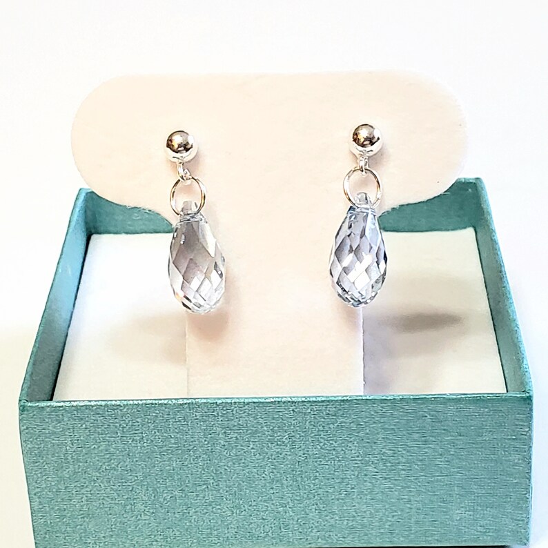 May include: A pair of silver-toned drop earrings with faceted, teardrop-shaped clear crystal pendants. The earrings are displayed on a white card inside a teal gift box, creating a visually appealing presentation. The earrings are a classic jewelry item.