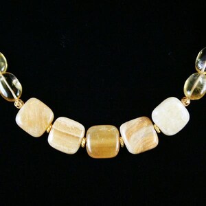 May include: A necklace featuring square, light-colored gemstones with gold-colored accents. The stones vary in shades of yellow and cream, with some displaying subtle patterns. The background is solid black.