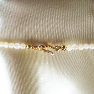 May include: Close-up of a delicate necklace featuring small, round, white beads. The necklace is secured with a gold-colored clasp. The background is a light, textured fabric, providing a soft contrast to the jewelry.