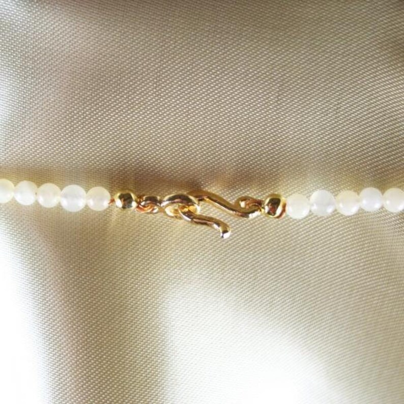 May include: Close-up of a delicate necklace featuring small, round, white beads. The necklace is secured with a gold-colored clasp. The background is a light, textured fabric, providing a soft contrast to the jewelry.
