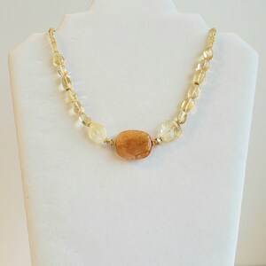 May include: A necklace featuring a large, rectangular, amber-colored stone as the focal point, surrounded by smaller, light yellow and clear beads. The beads are strung on a gold-colored chain, creating a delicate and elegant piece of jewelry.