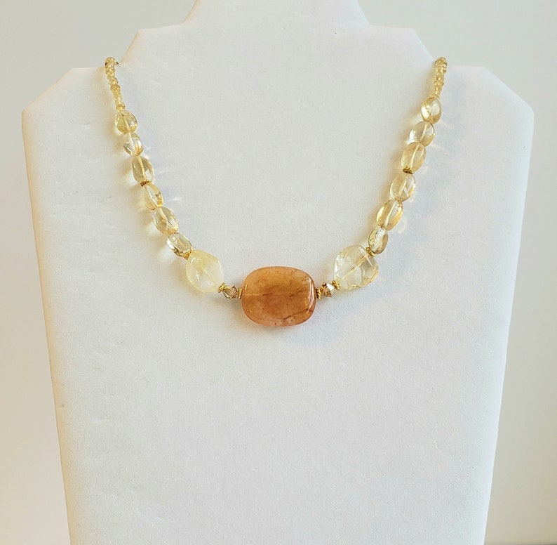 May include: A necklace featuring a large, rectangular, amber-colored stone as the focal point, surrounded by smaller, light yellow and clear beads. The beads are strung on a gold-colored chain, creating a delicate and elegant piece of jewelry.