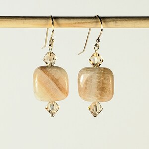 May include: A pair of dangle earrings featuring square, light-colored stone beads with a striped pattern. Each earring has a crystal accent above and below the stone, and gold-colored ear wires.