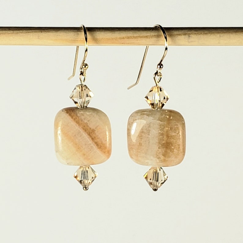 May include: A pair of dangle earrings featuring square, light-colored stone beads with a striped pattern. Each earring has a crystal accent above and below the stone, and gold-colored ear wires.