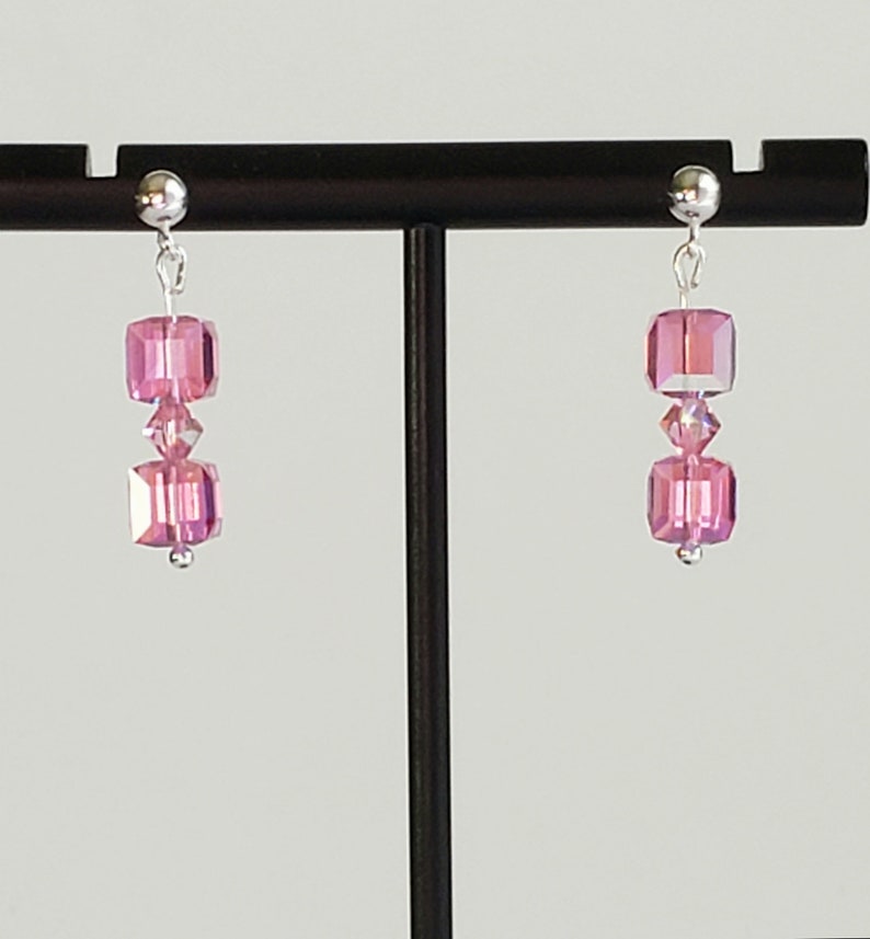 May include: A pair of silver earrings with pink crystal beads. The earrings are hanging from a black stand.