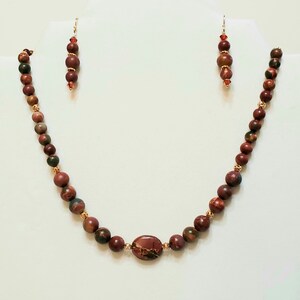 May include: A red, brown, and green stone beaded necklace with a larger oval-shaped stone pendant. The necklace is paired with matching earrings.
