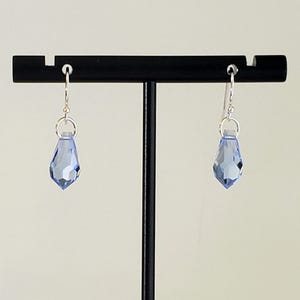 May include: A pair of light blue teardrop-shaped earrings with faceted surfaces. The earrings are suspended from silver-colored hooks and are displayed on a black stand against a neutral background. The earrings are a delicate accessory.