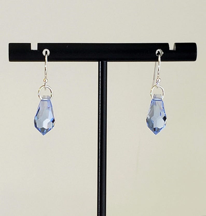 May include: A pair of light blue teardrop-shaped earrings with faceted surfaces. The earrings are suspended from silver-colored hooks and are displayed on a black stand against a neutral background. The earrings are a delicate accessory.