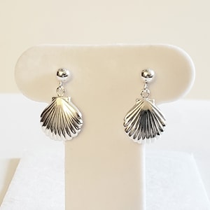 May include: A pair of silver seashell earrings. Each earring features a small, round stud connected to a detailed, fan-shaped seashell pendant. The earrings are displayed on a white surface against a neutral background.