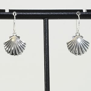 May include: A pair of silver earrings with a seashell design. The earrings are hanging from a black display stand.