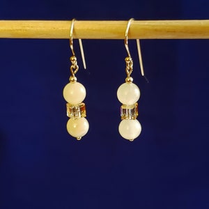 May include: A pair of dangle earrings with a gold-colored hook. Each earring features two off-white, round beads separated by a square, iridescent crystal bead. The earrings are displayed against a dark blue background.