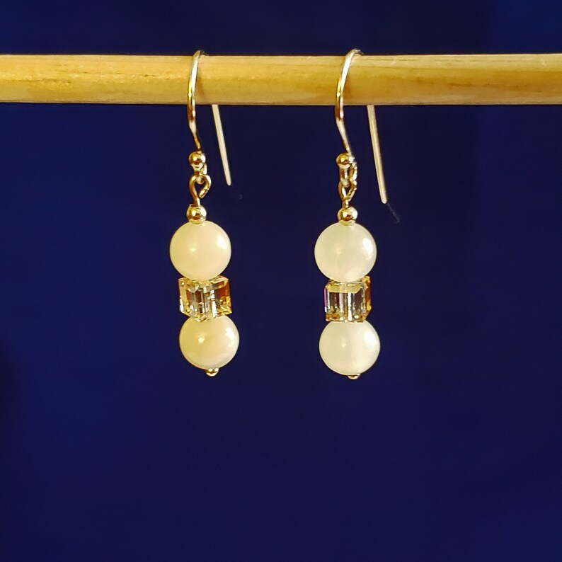May include: A pair of dangle earrings with a gold-colored hook. Each earring features two off-white, round beads separated by a square, iridescent crystal bead. The earrings are displayed against a dark blue background.