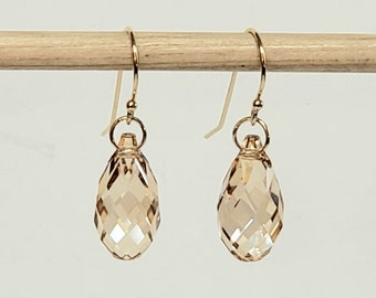 May include: A pair of drop earrings featuring faceted, champagne-colored teardrop crystals suspended from gold-toned metal ear wires. Each crystal is attached to a small circular gold-toned jump ring, creating a dangling design with light-reflecting surfaces.
